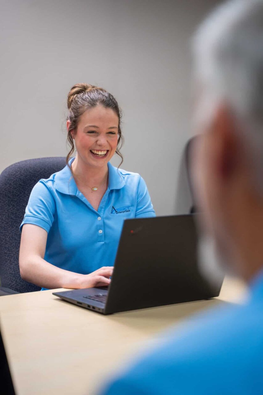 Smiling woman in blue Asset Enterprises polo using a laptop, representing streamlined IT support in retail technology.