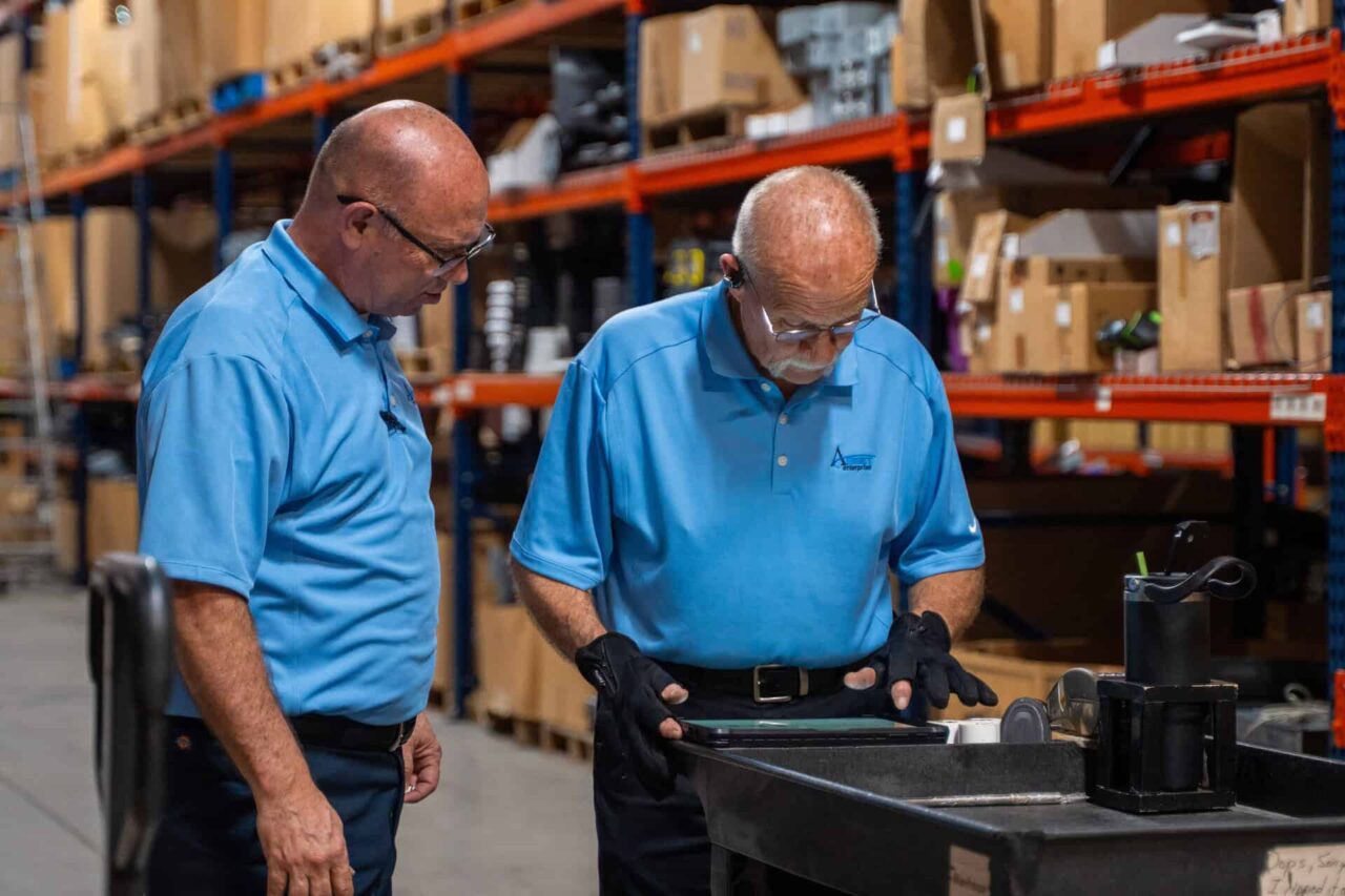 Two workers in blue shirts inspect equipment in a warehouse, ensuring control and consistency for retailers.